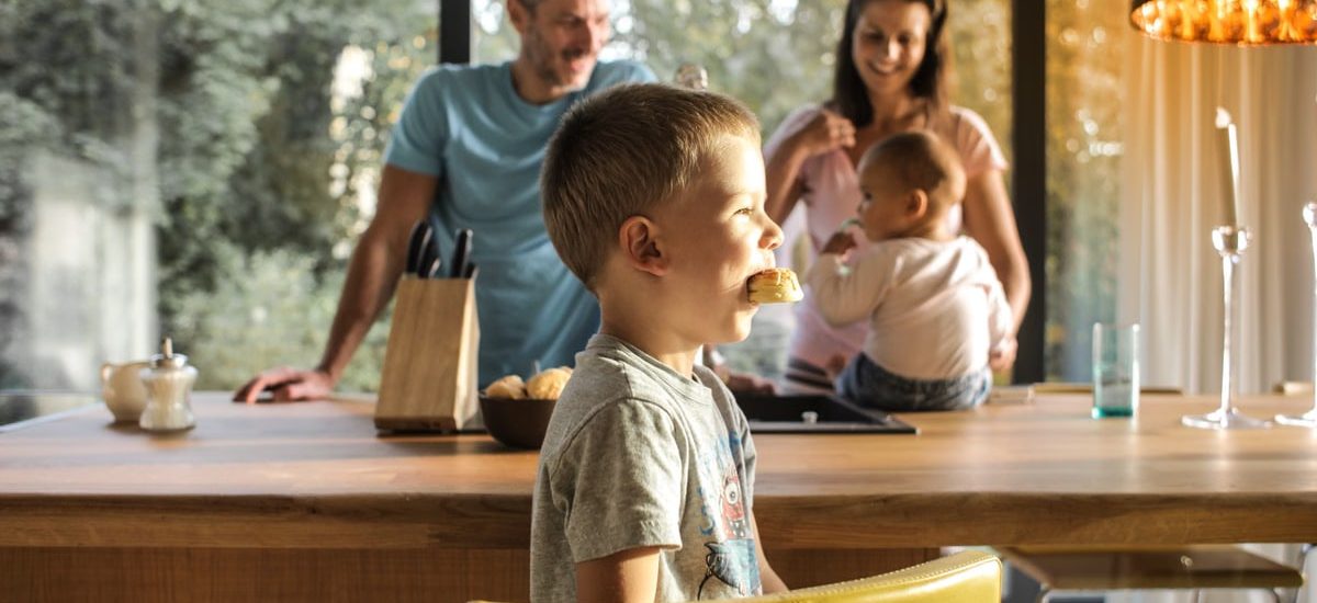 Side view of playful boy eating cookie while sitting at table with parents and sibling on background spending time at home