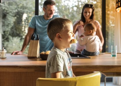 Side view of playful boy eating cookie while sitting at table with parents and sibling on background spending time at home